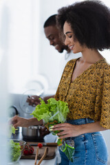 Side view of smiling african american woman holding lettuce near blurred boyfriend in kitchen.