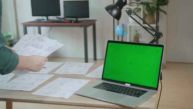 Creative Designer Works On A Storyboard, Looks At His Sketches With Mock Up Green Screen Laptop Computer, Choosing The Best Drawings For His Project, Comics Compilation, Application Ui Or Game Plot
