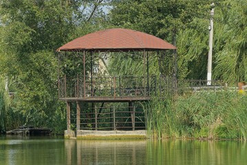 open brown empty gazebo with wrought iron fence on the shore above the water in green vegetation