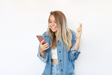 A young caucasian blonde woman in a denim jacket and jeans crosses her fingers for good luck looking at the phone screen waiting for the results of the lottery or exams isolated on a white background