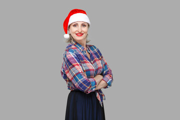 Portrait of confident attractive adult woman in red santa cap and checkered shirt standing and closed up hands with toothy smiling, looking at camera. Indoor, studio shot, yellow background