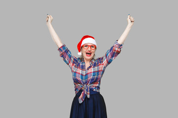 Fototapeta na wymiar Portrait of funny modern aged woman in red santa cap, and eyeglasses checkered shirt standing with hands up and with surprised face looking at camera. Indoor, studio shot, yellow background