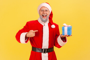 Extremely happy elderly man with gray beard wearing santa claus costume holding present box and pointing at gift with positive look. Indoor studio shot isolated on yellow background.