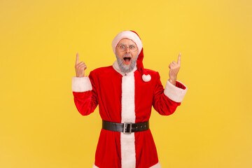 Fototapeta na wymiar Excited screaming elderly man with gray beard in santa claus costume pointing up with both hands, keeps mouth open, presenting copy space above head. Indoor studio shot isolated on yellow background.