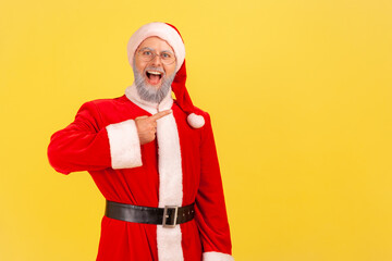 Positive excited elderly man with gray beard wearing santa claus costume with open mouth and excited expression, pointing at copy space. Indoor studio shot isolated on yellow background.