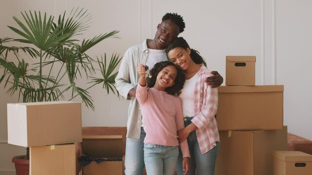 Happy young african american mother, father and daughter smiling to camera, girl showing keys, posing in new home