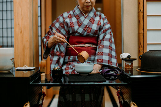 Woman In Red And Blue Kimono Holding Standing Beside A Table Pouring Clear Liquid In A Ceramic Bowl