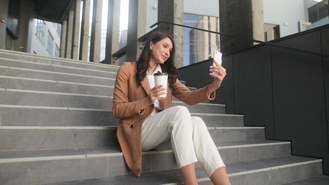 Caucasian young attractive business lady sitting on stairs outdoors near office centre and have a video call. Woman talking on video conference online outdoors. High quality 4k footage