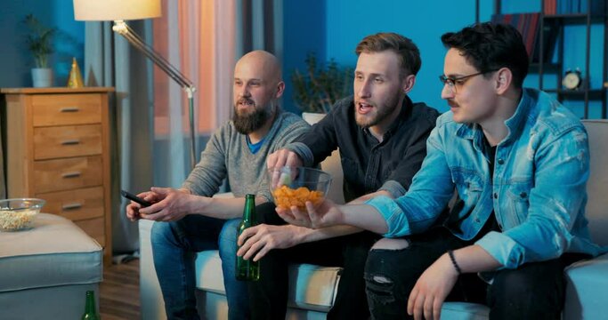 Men's evening in front of TV on sofa, guys drinking beer, eating popcorn chips talking watching game on TV, cheering for team, shouting, reliving goal, raising their hands clenched into fists, victory