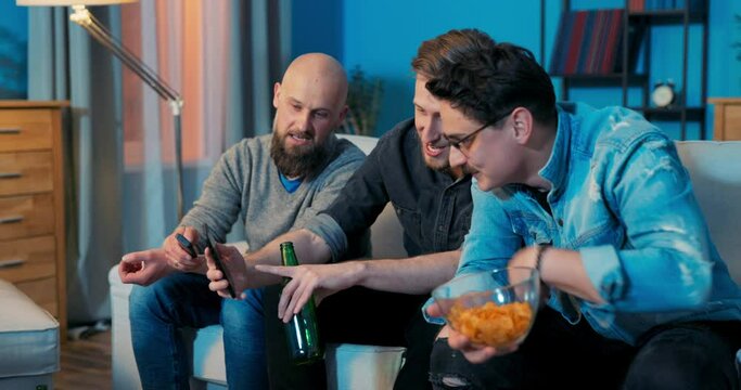 Man shows friends on phone pictures of his new girlfriend, text message, they sit together on couch, talk, advise, comment, drink beer, eat chips, watch TV, men's night out