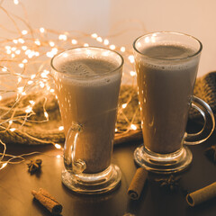 Couple of hot cocoa drinks in glasses on brown table. Hot chocolate. Knitted scarf. Cloves, cinnamon and star anise. Led lights garland.