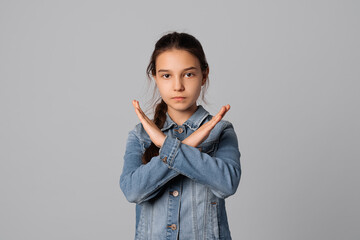 Serious young girl making X sign with her arms to stop doing something, standing over grey...