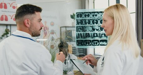 Medicine concept where good-looking smart qualified adult male and female medics standing near glass wall in workroom and discussing results of x-ray scan and drawing chemical formula on this wall - Powered by Adobe