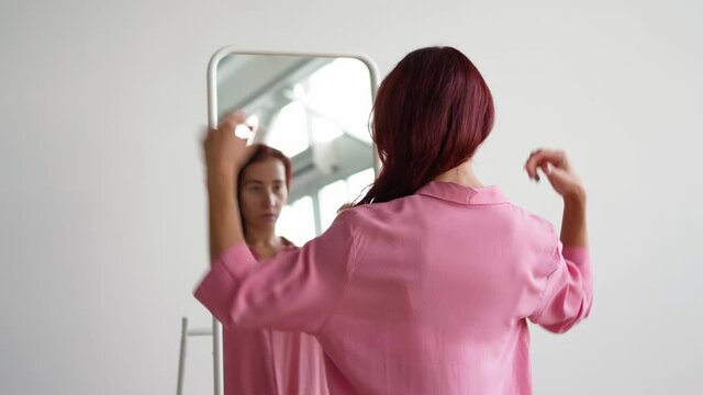 Young woman in a pink shirt straightens her red hair near the mirror, getting ready for work in the morning. 