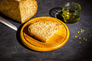 Slice of rustic natural yeast-free bread with flax, poppy seeds, sesame seeds, millet, pumpkin and sunflower seeds, with olive oil in a glass jar, and a knife, on a wooden board, black background