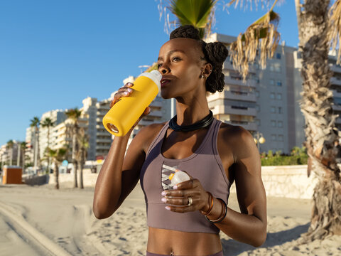 Black sportswoman drinking water on beach