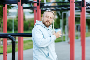 A man with a braid hairstyle in the early morning drinks coffee on the sports ground and listens to music on headphones.