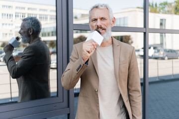 bearded journalist in blazer holding microphone and making reportage near building