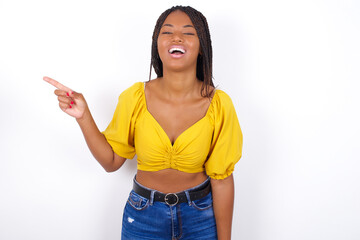 Young african american woman with braids over white wall laughs happily points away on blank space demonstrates shopping discount offer, excited by good news or unexpected sale.