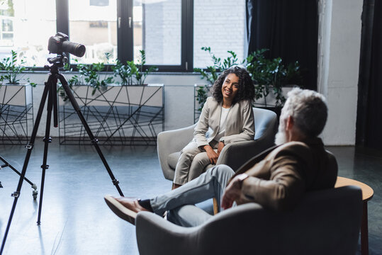 happy african american journalist and blurred businessman talking during interview near digital camera on tripod