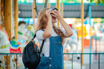 A young woman tourist in a denim sundress stands with cameras in an amusement park.