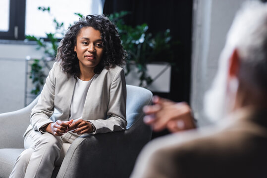 pretty african american journalist looking at blurred businessman sitting in armchair and gesturing while talking during interview