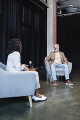 Obraz premium curly african american journalist in suit talking with bearded businessman sitting in armchair during interview