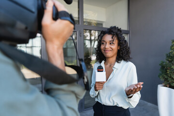 happy african american journalist with microphone gesturing while doing reportage near blurred...