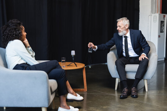 businessman in suit taking glass of water during interview near african american journalist