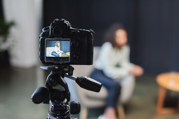 curly african american journalist sitting in armchair near blurred digital camera