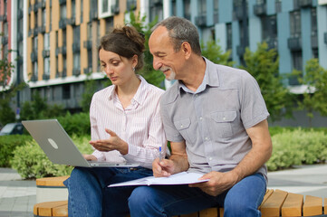 Attractive young woman and senior man are using laptop sitting on the bench in modern residential complex. Casual meeting outside office. A daughter is teaching technology for her father