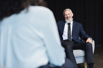 cheerful businessman in suit looking at blurred journalist during interview