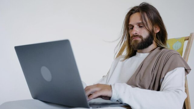 Jesus sits in a schizlong and works at a computer on a white background. Typing on the computer. Studio. High quality 4k footage