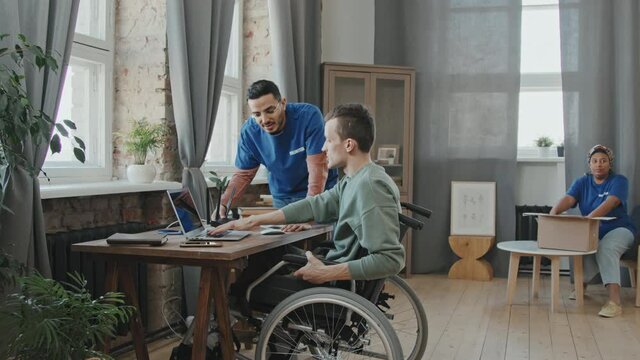 Zoom-in slowmo shot of group of volunteers helping young man with disability at his apartment, setting up internet on laptop computer and unpacking clothes from cardboard boxes
