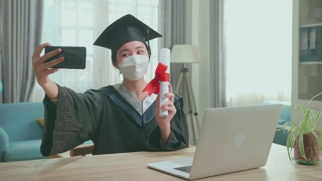 Young Asian Woman Wearing Protection Face Mask, Taking A Selfie While Wearing A Graduation Gown And Cap, Hand Holding University Certificate In Living Room
