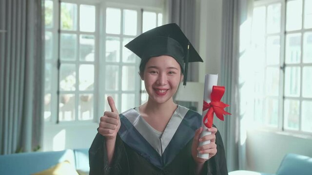 Excited Asian Woman Holding A University Certificate, Smiling And Thumb Up To Camera. Pretty Female Graduate Wearing A Graduation Gown And Cap Sitting On The Living Room
