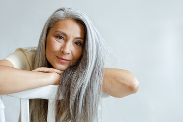 Cheerful middle aged Asian lady with flowing hoary hair sits backwards on chair on light background...