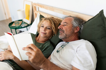 Joyful senior couple reading a book together on the bed