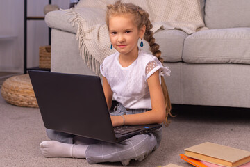 An elementary school girl sits at home on the couch with a laptop in an online lesson during quarantine due to the coronavirus pandemic.