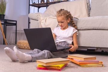 An elementary school girl sits at home on the couch with a laptop in an online lesson during quarantine due to the coronavirus pandemic.