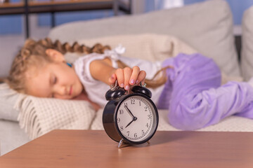 Little girl sleeping in bed with alarm clock on the table. Wake up early in the morning. We put the...