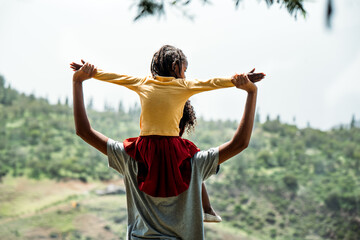 black mother holding on shoulders black little daughter on top of mountain