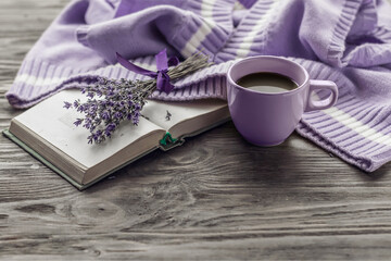Morning coffee. A cup of coffee on a wooden table, an open book and a warm sweater against the background of a bouquet of flowers. Still life in lavender color. Cozy morning.