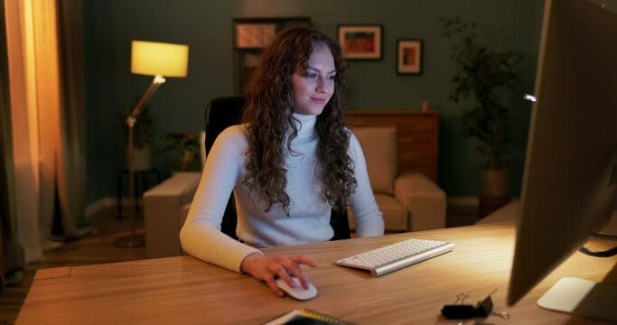 A teenager spends evenings in front of the computer, scrolling through websites, scrolling through pictures on social media with mouse, smiling as she looks at her friends' posts, laughing