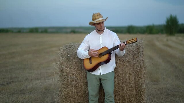man wearing a straw hat playing the guitar about a haystack