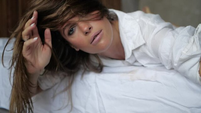 attractive model lies on a white bed and straightens her hair, looking directly into the camera. High quality FullHD footage