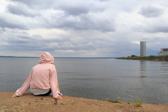 Female in a pink hood sweater looking out over the water. One gray factory or industry in the background. Part of the Swedish lake called Vänern or Vanern. Lidköping, Sweden, Europe.