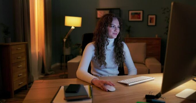 A teenager spends evenings in front of the computer, scrolling through websites, scrolling through pictures on social media with mouse, smiling as she looks at her friends' posts, laughing