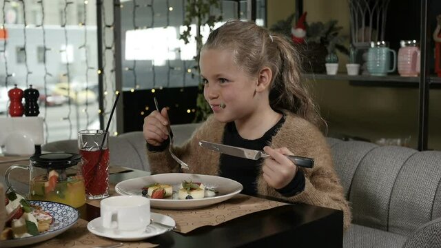 cute little girl eats cheesecakes with fruit and smiles sitting on a gray sofa in a restaurant. High quality FullHD footage