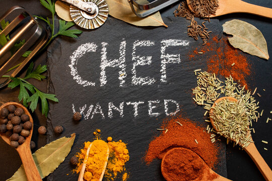 Chef wanted written on black stone tile with white chalk, surrounded by condiments an kitchen tools (paprika, curcuma, all spice, parsley, wood spoons...)
